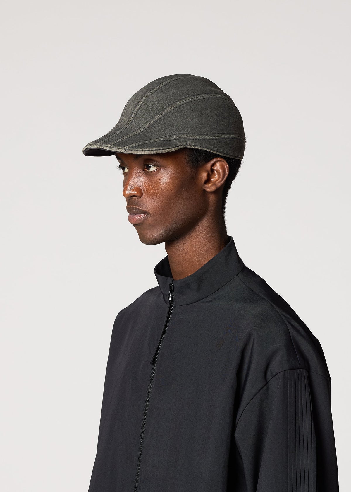 A person wearing the IM MEN CARVED CAP, a dark adjustable cap with a three-dimensional pattern, and a black zip-up jacket stands against a plain light background facing left. view 5 of 6