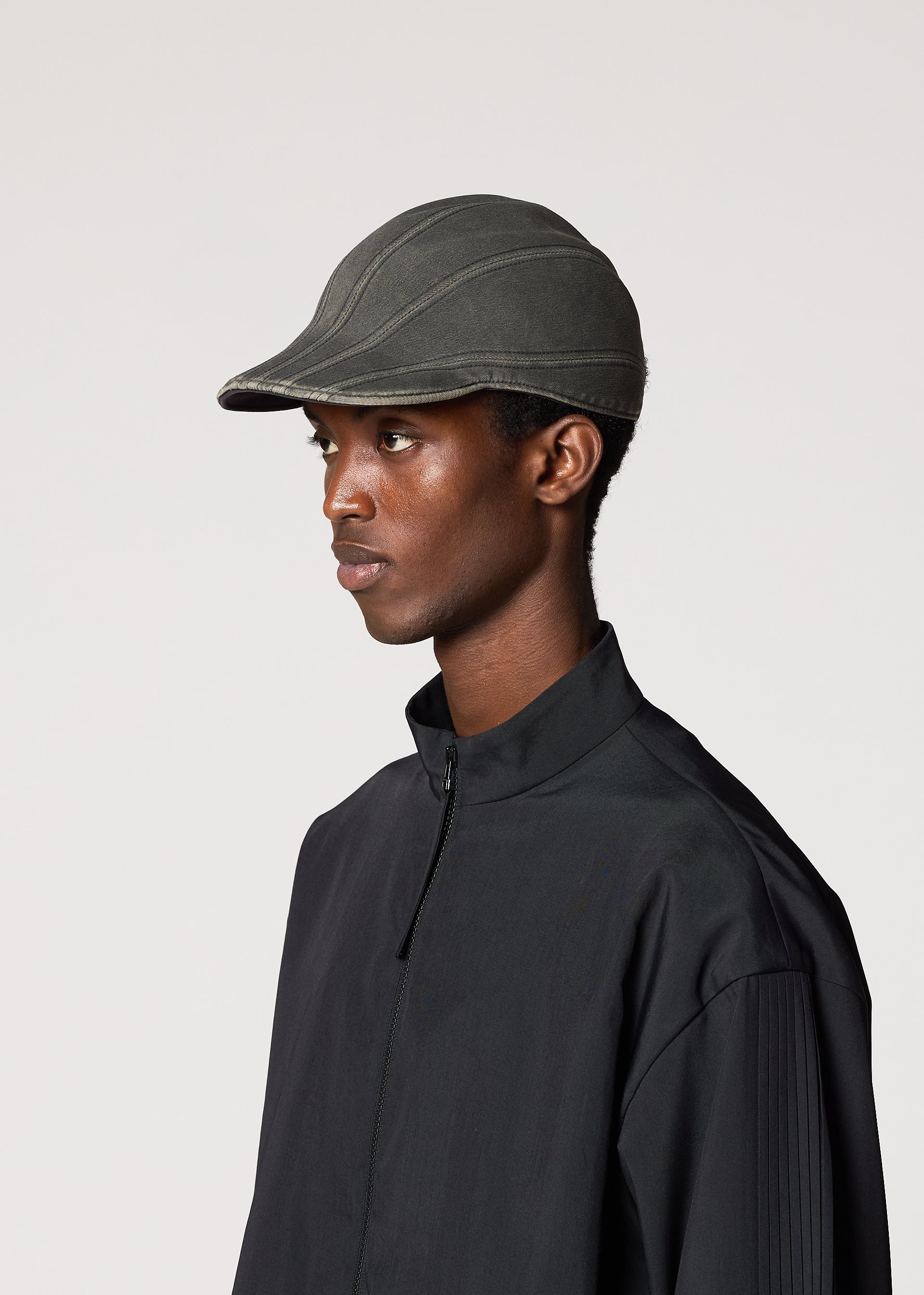 Zoomed view A person wearing the IM MEN CARVED CAP, a dark adjustable cap with a three-dimensional pattern, and a black zip-up jacket stands against a plain light background facing left.