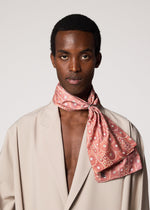 A person stands against a plain light background, wearing an IM MEN UROKOMON STOLE—a pink, scale-patterned silk scarf—tied around their neck with a beige blazer and no shirt underneath.