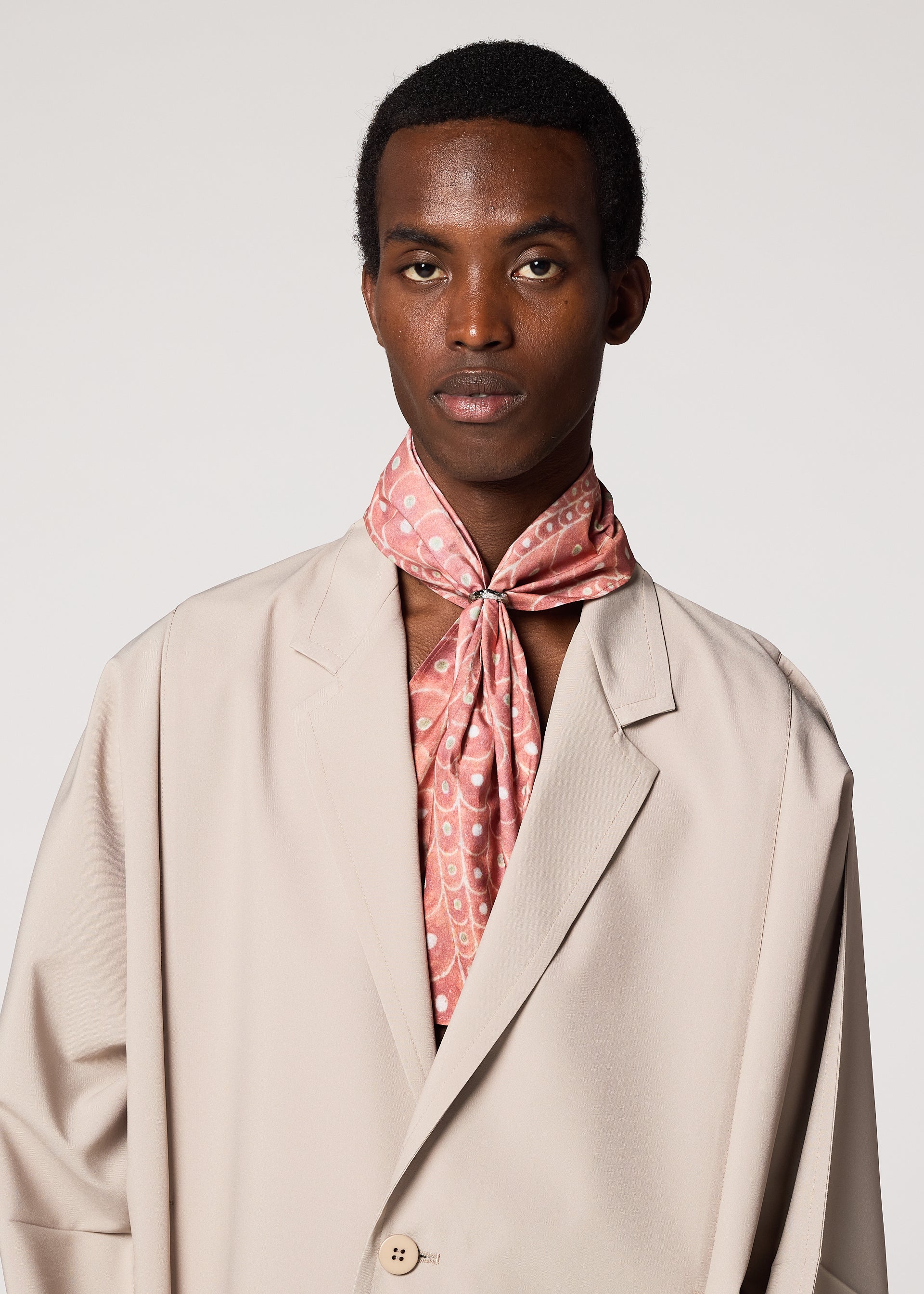 Zoomed view A person wearing the IM MEN UROKOMON STOLE—featuring a light pink, scale-like cotton design—neatly tied around their neck with a beige blazer, standing against a plain white background.
