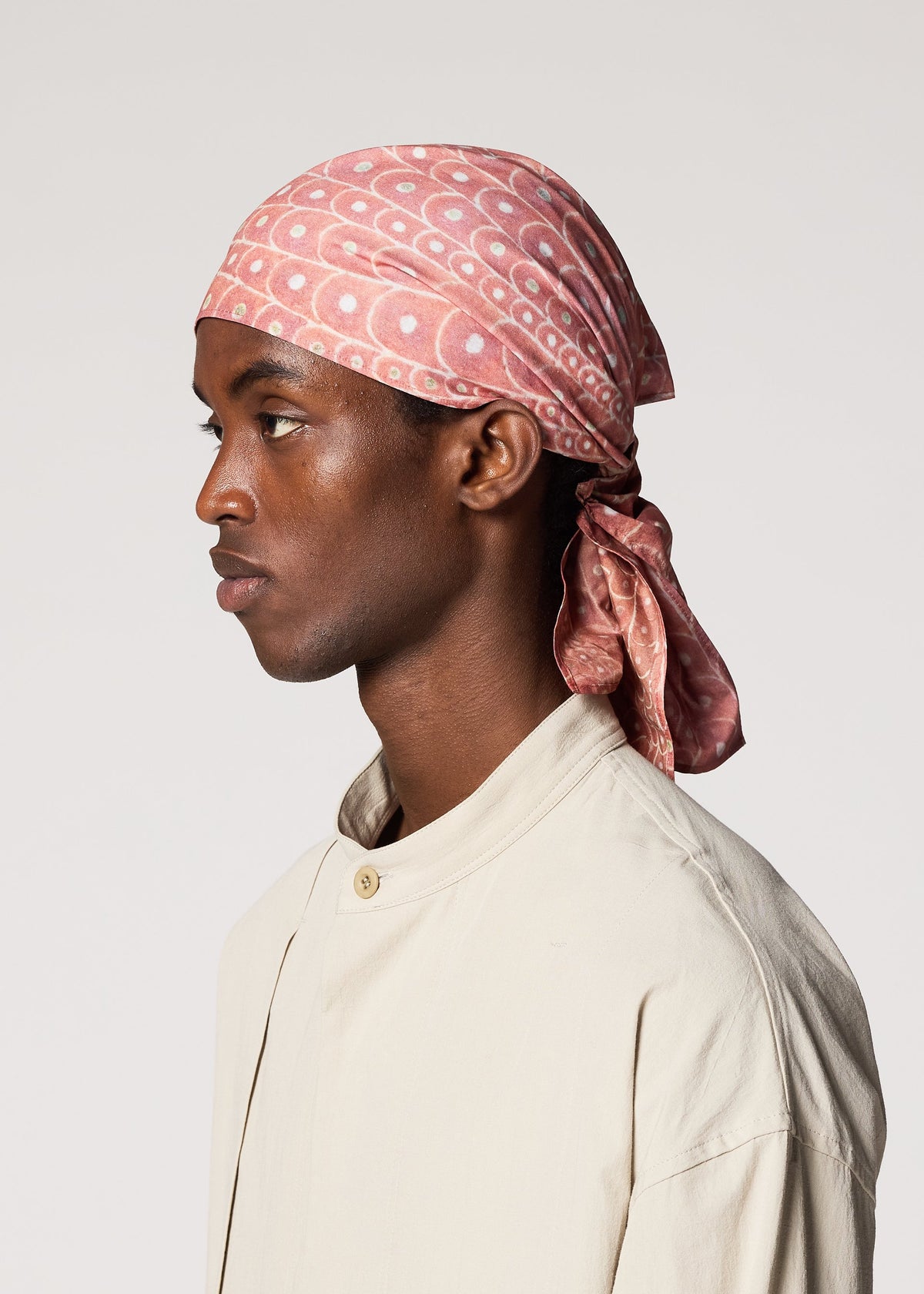 A person in profile wears the IM MEN UROKOMON STOLE, a pink patterned cotton accessory with a subtle scale-like design, paired with a light beige shirt against a plain light background. view 5 of 5