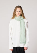 A person with wavy, shoulder-length blond hair wears a loose white button-up shirt and the IM MEN SHRINK BORDER STOLE in light green linen, standing against a plain light background.