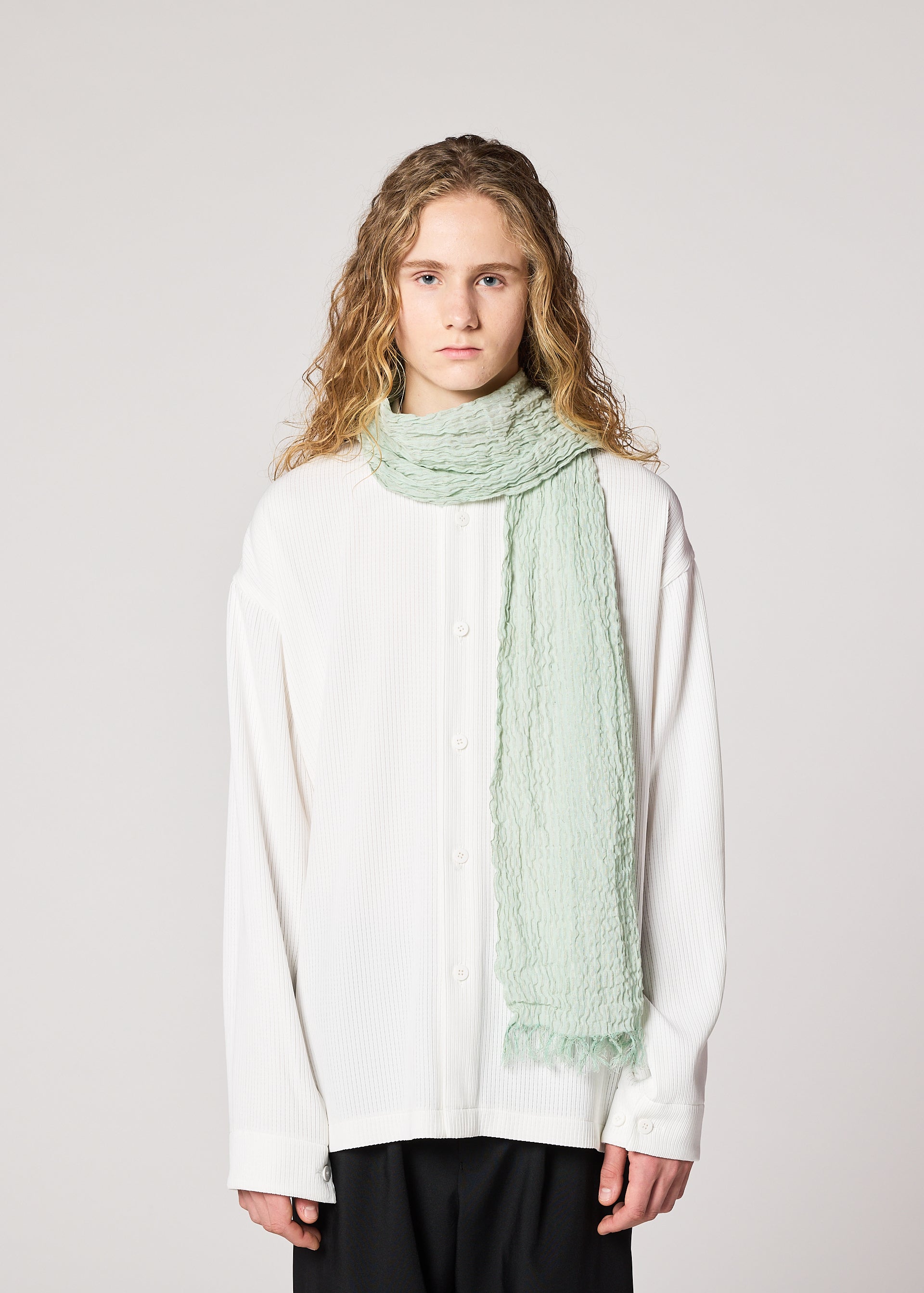 Zoomed view A person with wavy, shoulder-length blond hair wears a loose white button-up shirt and the IM MEN SHRINK BORDER STOLE in light green linen, standing against a plain light background.