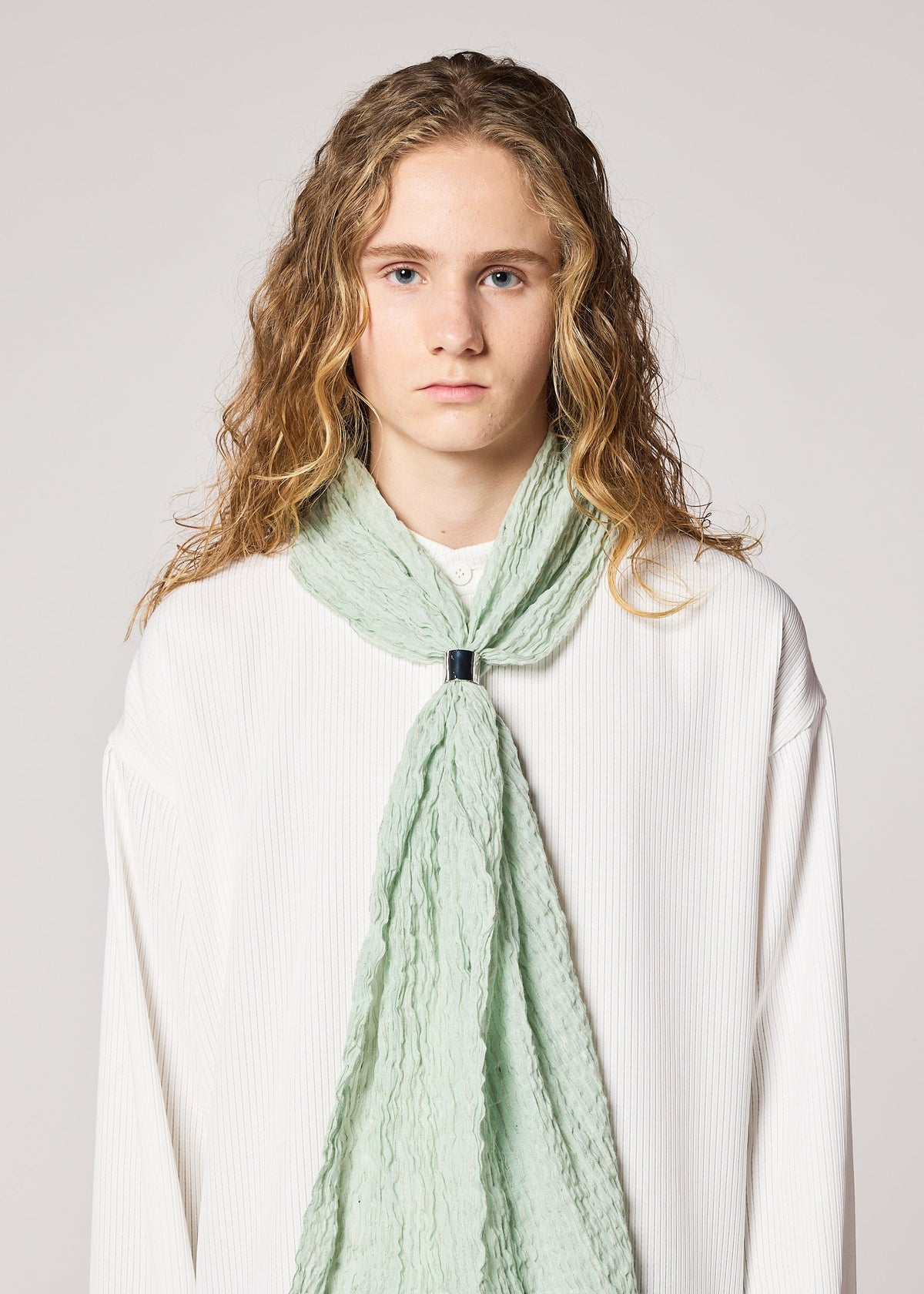 A person with long, wavy blonde hair wears the breathable pale green "SHRINK BORDER STOLE" by IM MEN, fastened with a black clasp over a loose-fitting white ribbed top, standing against a plain light gray background. view 6 of 7