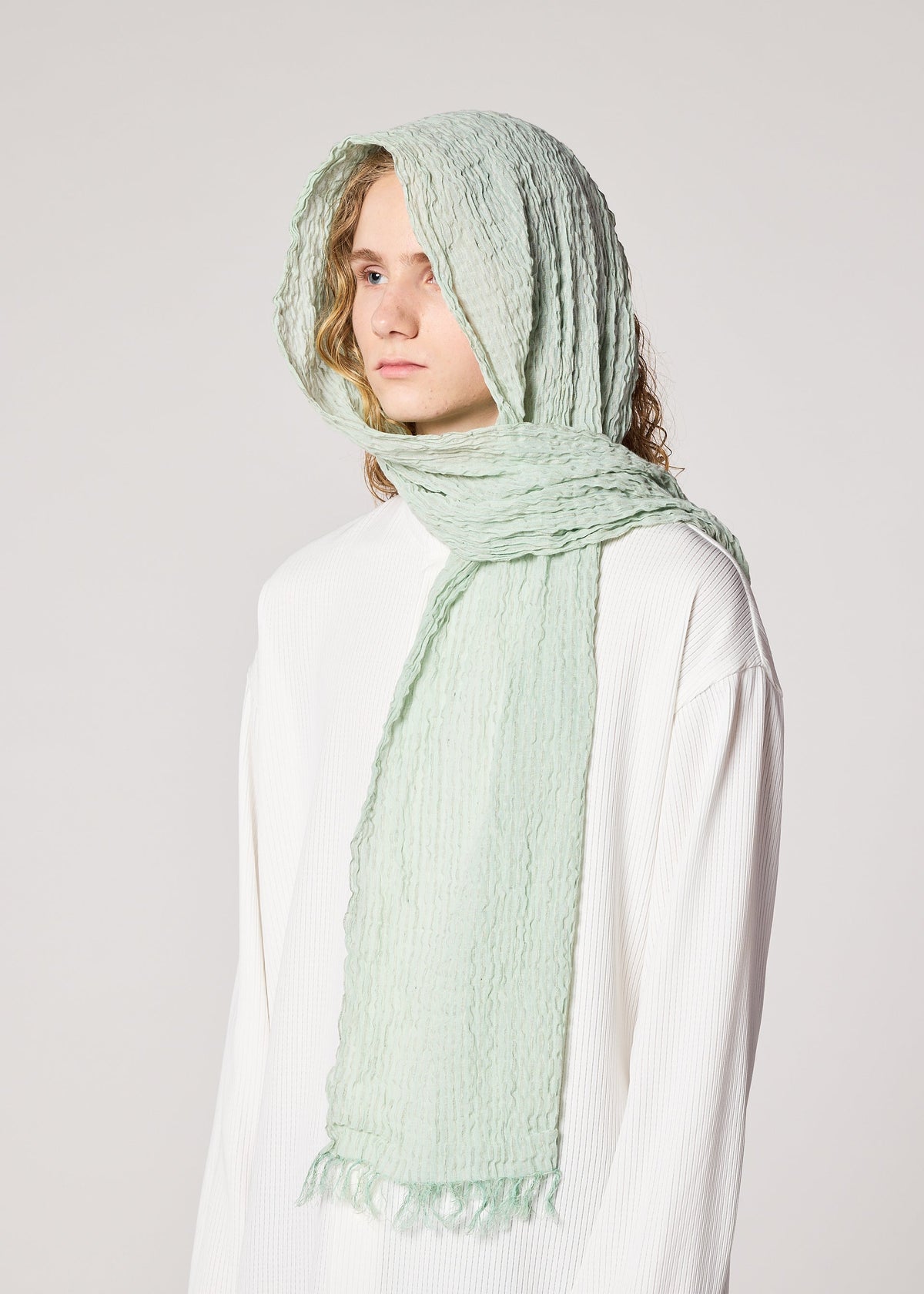 A person wearing the IM MEN SHRINK BORDER STOLE in light mint green, draped over their head and neck, paired with a loose white high-twist cotton top, stands against a plain light background. view 7 of 7
