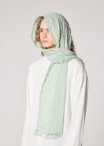 A person wearing the IM MEN SHRINK BORDER STOLE in light mint green, draped over their head and neck, paired with a loose white high-twist cotton top, stands against a plain light background.