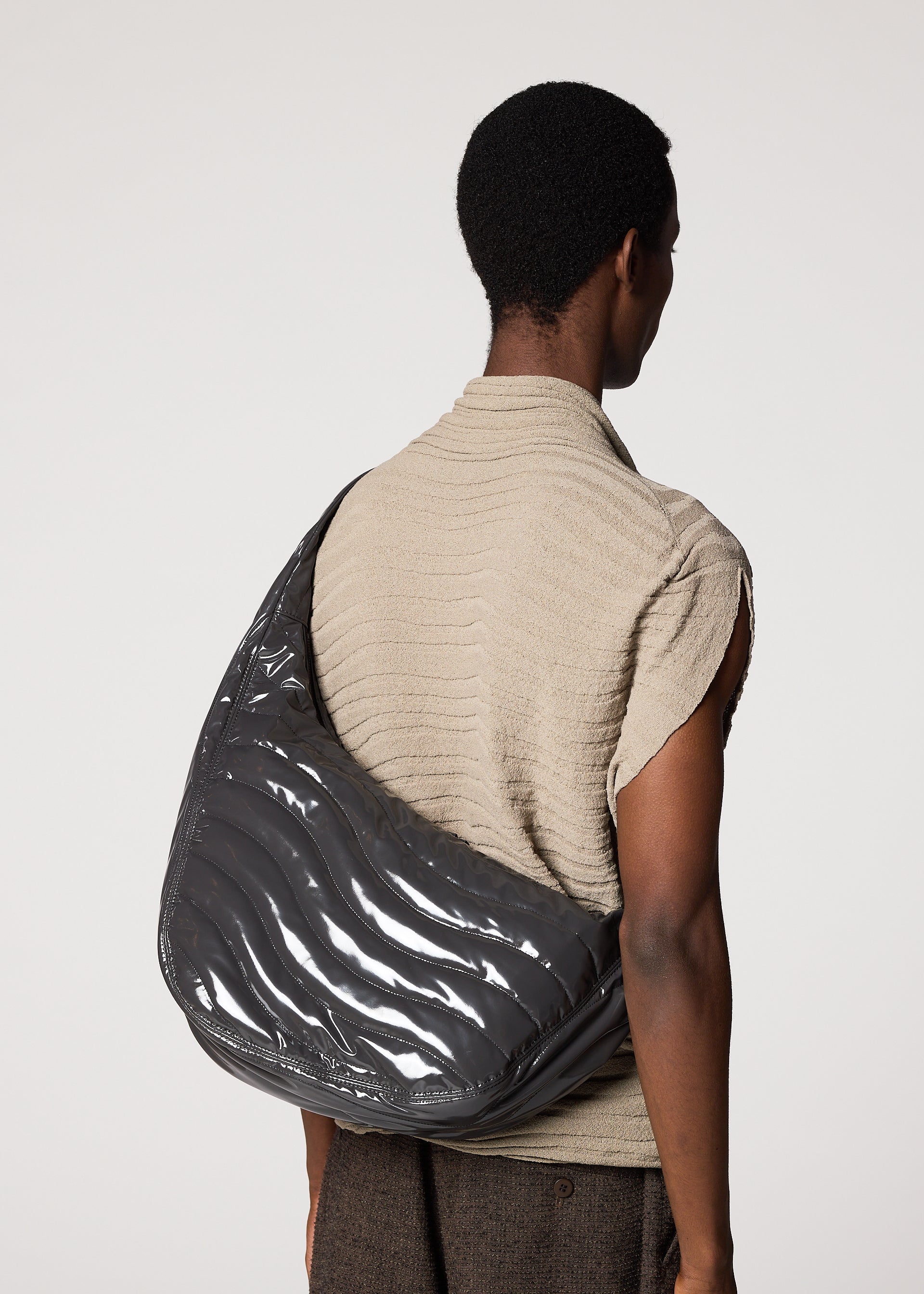 Zoomed view A person with short hair, seen from behind, wears a textured beige top and carries the IM MEN ENGRAVE BAG—a large, sculptural bag featuring waved patterns and an adjustable shoulder strap—against a plain light background.