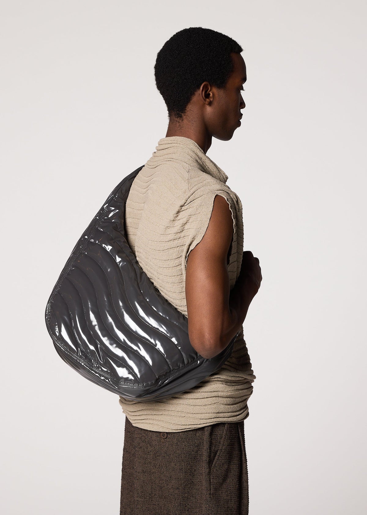 A person in a textured, sleeveless beige top and brown pants stands in profile, carrying the IM MEN ENGRAVE BAG—a sculptural accessory featuring waved patterns and an adjustable shoulder strap—against a plain background. view 6 of 6