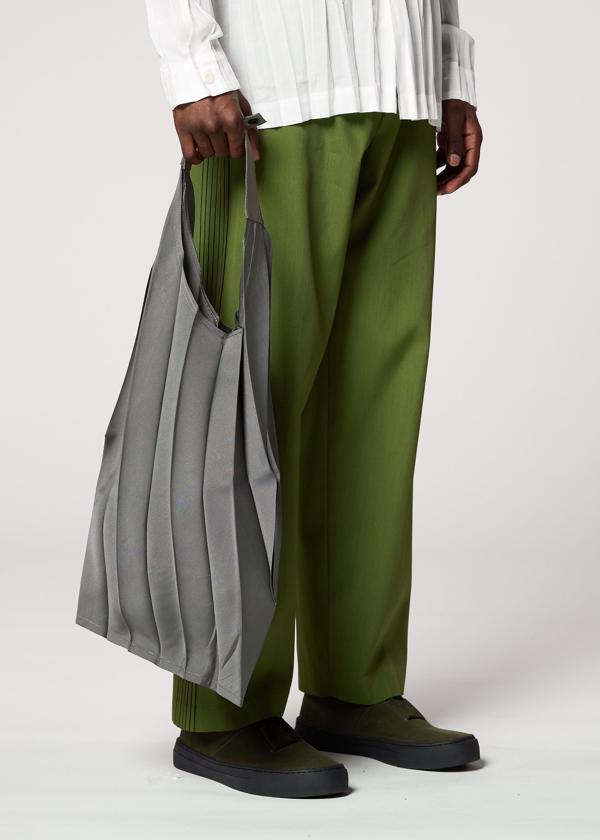 Zoomed view A person dressed in green pants, a white pleated shirt, and dark green shoes holds the gray GURU GURU foldable tote bag with handmade pleats by IM MEN at their side against a plain background.