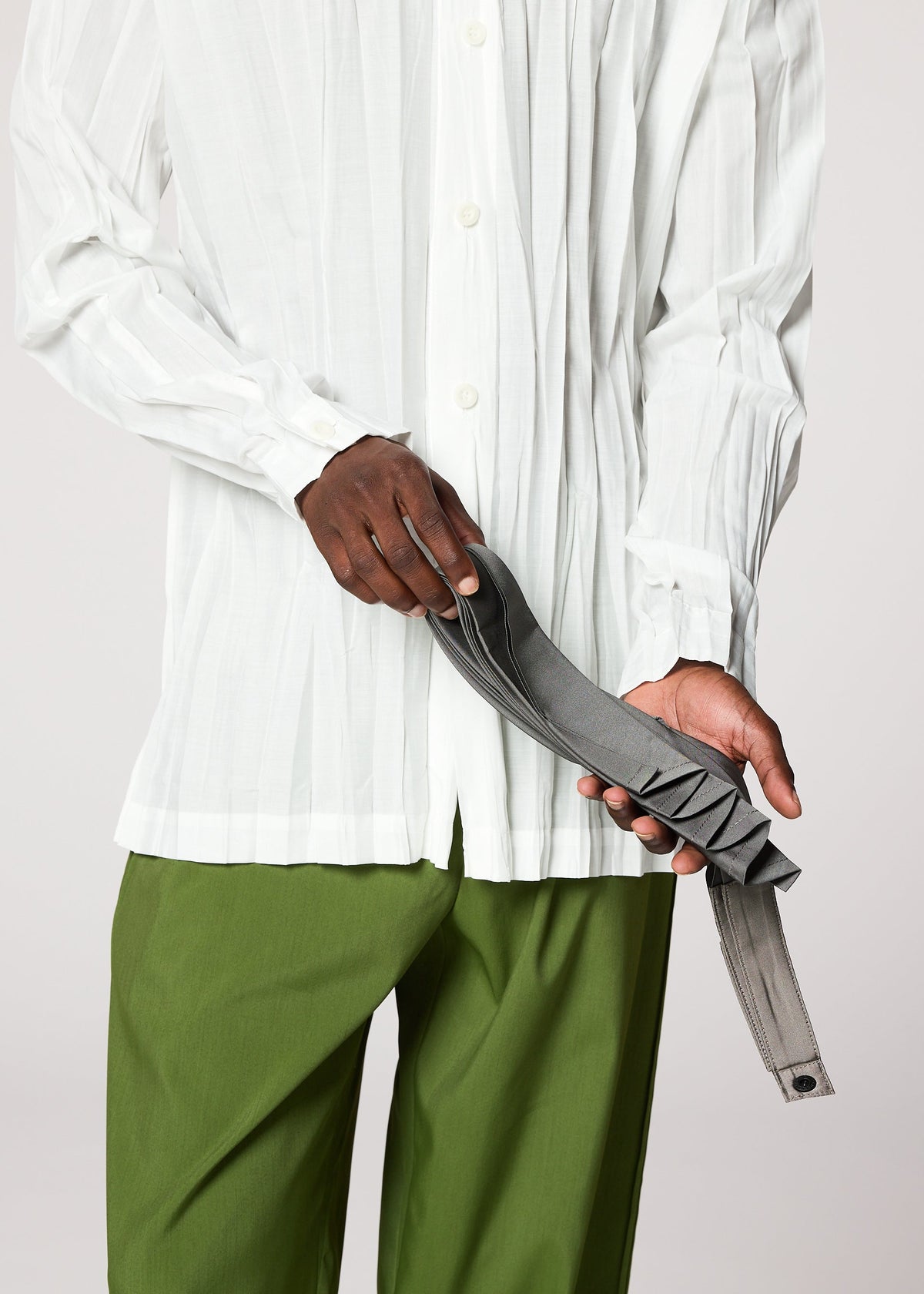 Someone in a white button-up shirt and green pants holds the gray handmade-pleated GURU GURU belt by IM MEN against a plain background. view 9 of 10