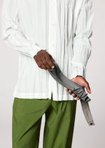 Someone in a white button-up shirt and green pants holds the gray handmade-pleated GURU GURU belt by IM MEN against a plain background.