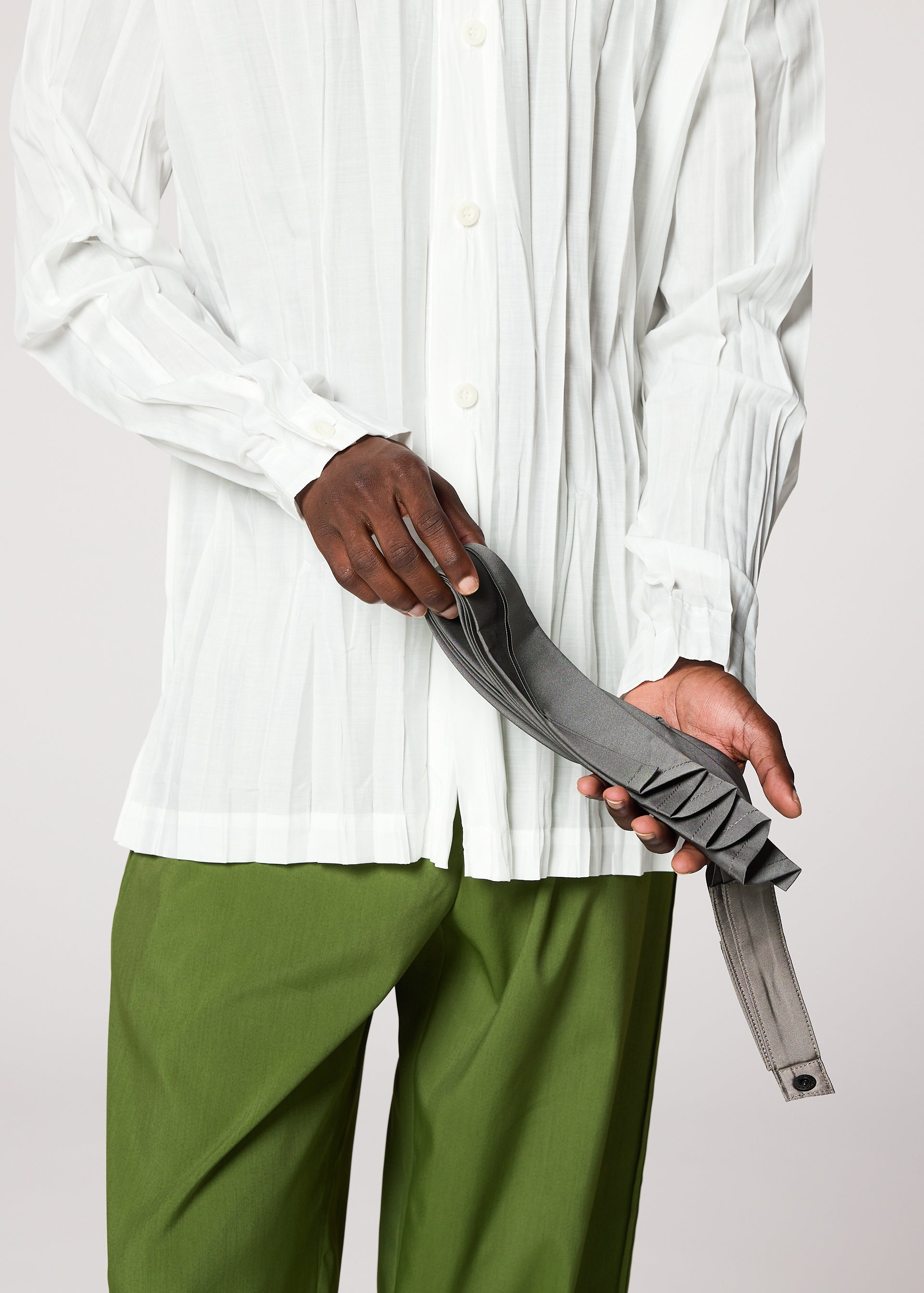 Zoomed view Someone in a white button-up shirt and green pants holds the gray handmade-pleated GURU GURU belt by IM MEN against a plain background.