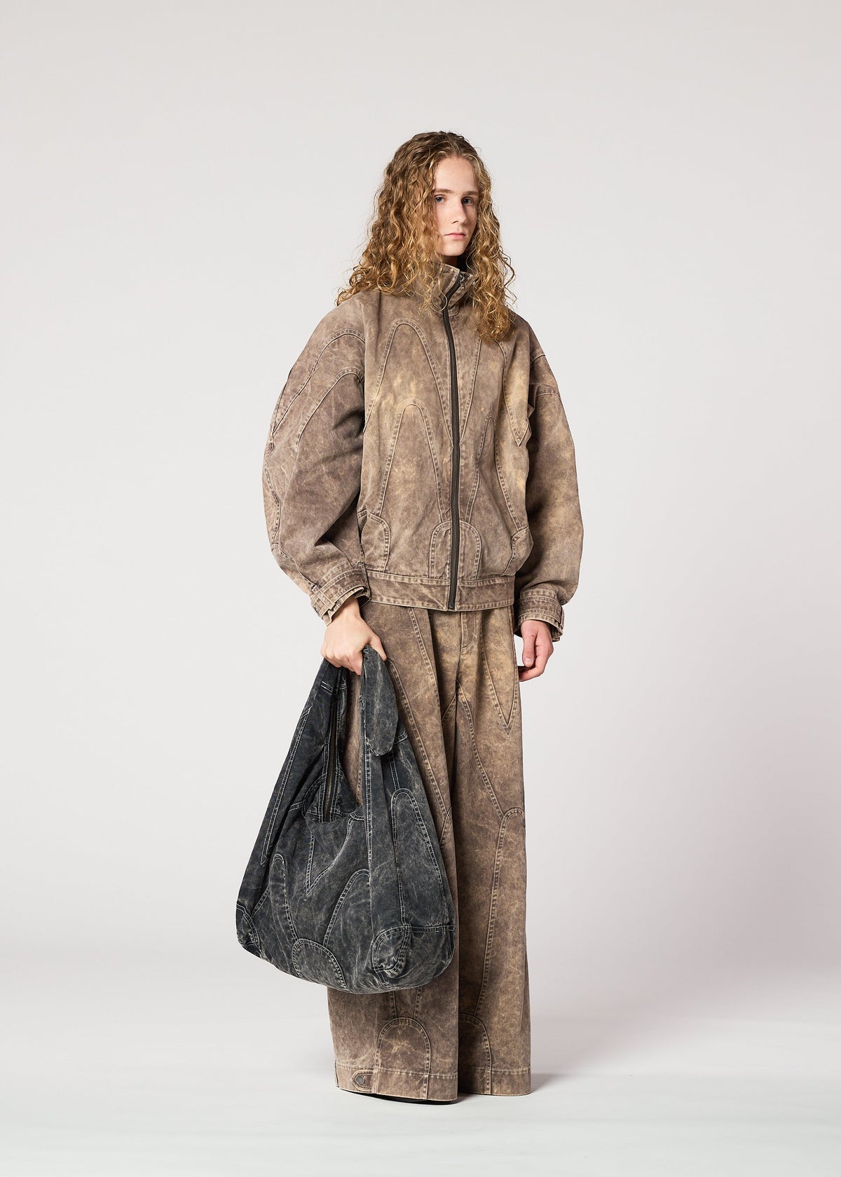 A person with long curly hair wears a loose, brown vintage-style zip-up jacket and wide-leg pants, holding the IM MEN ELEMENTS BAG—a large, ceramic-inspired black handbag—against a plain light background. view 4 of 5