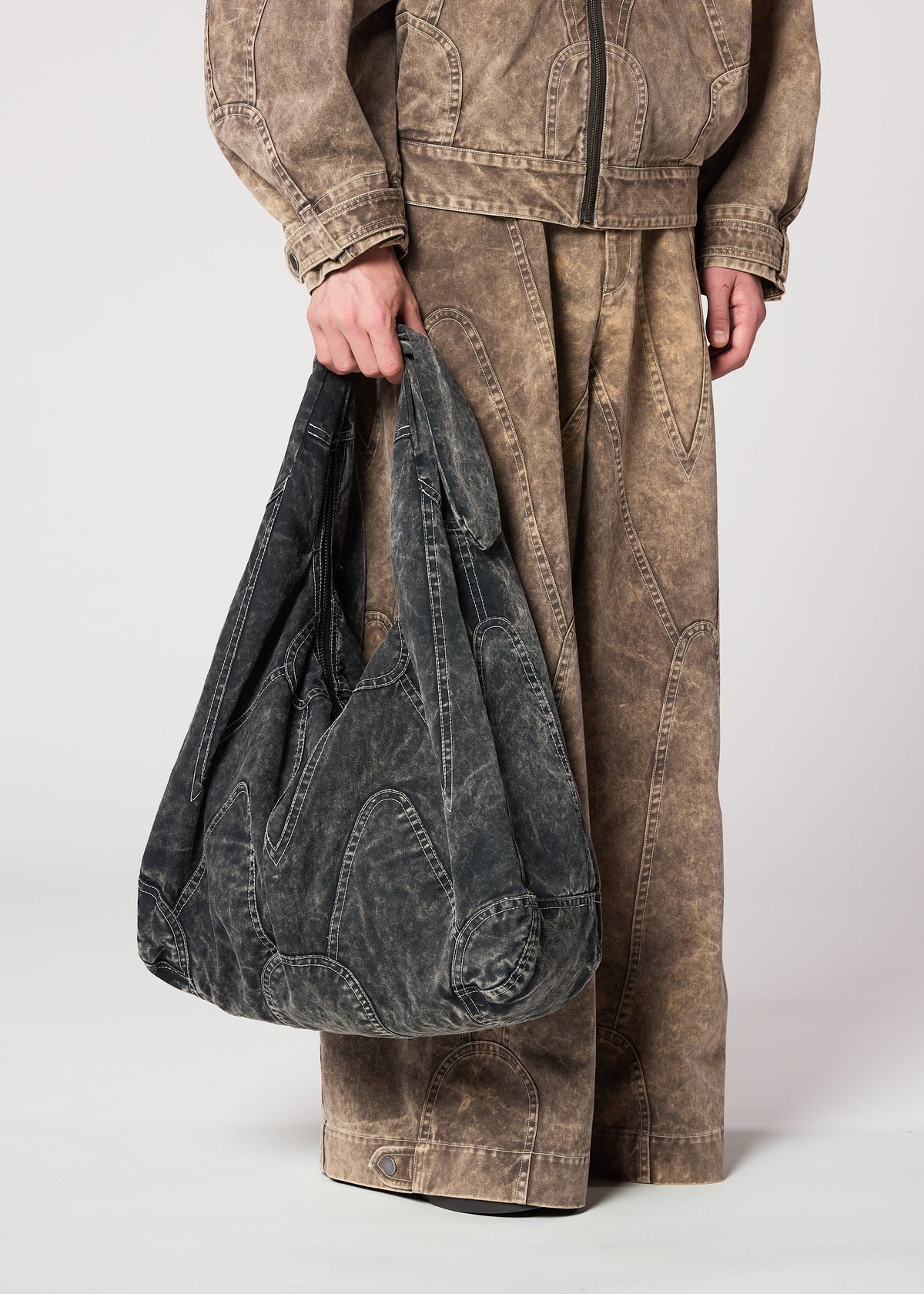 The lower half of a person in matching brown denim jacket and wide-leg pants is shown holding the IM MEN ELEMENTS BAG, a large black denim tote with ceramic-inspired design and visible seams, against a plain background. view 5 of 5