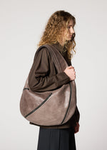 A person with long curly hair in a brown shirt and dark pants stands in profile, carrying the IM MEN CARVED LEATHER BAG with multiple zippers and an adjustable strap over their shoulder against a plain white background.