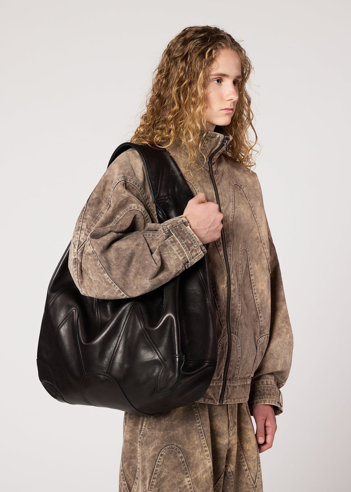 A person with long curly hair wears a loose, faded brown jacket and matching pants, holding the IM MEN ELEMENTS LEATHER BAG with an adjustable shoulder strap over one shoulder, standing against a plain white background. view 4 of 5