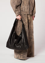 A person in a loose, brown, acid-wash denim jacket and wide-leg pants holds the IM MEN ELEMENTS LEATHER BAG by its handle against a plain background. The luxury bag features an adjustable shoulder strap.