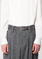 A person wearing IM MEN’s OVAL one-size-fits-most natural leather belt with an adjustable buckle, styled over gray textured trousers and a white button-up shirt. The photo is cropped at the shoulders and mid-thigh, hands at their sides.
