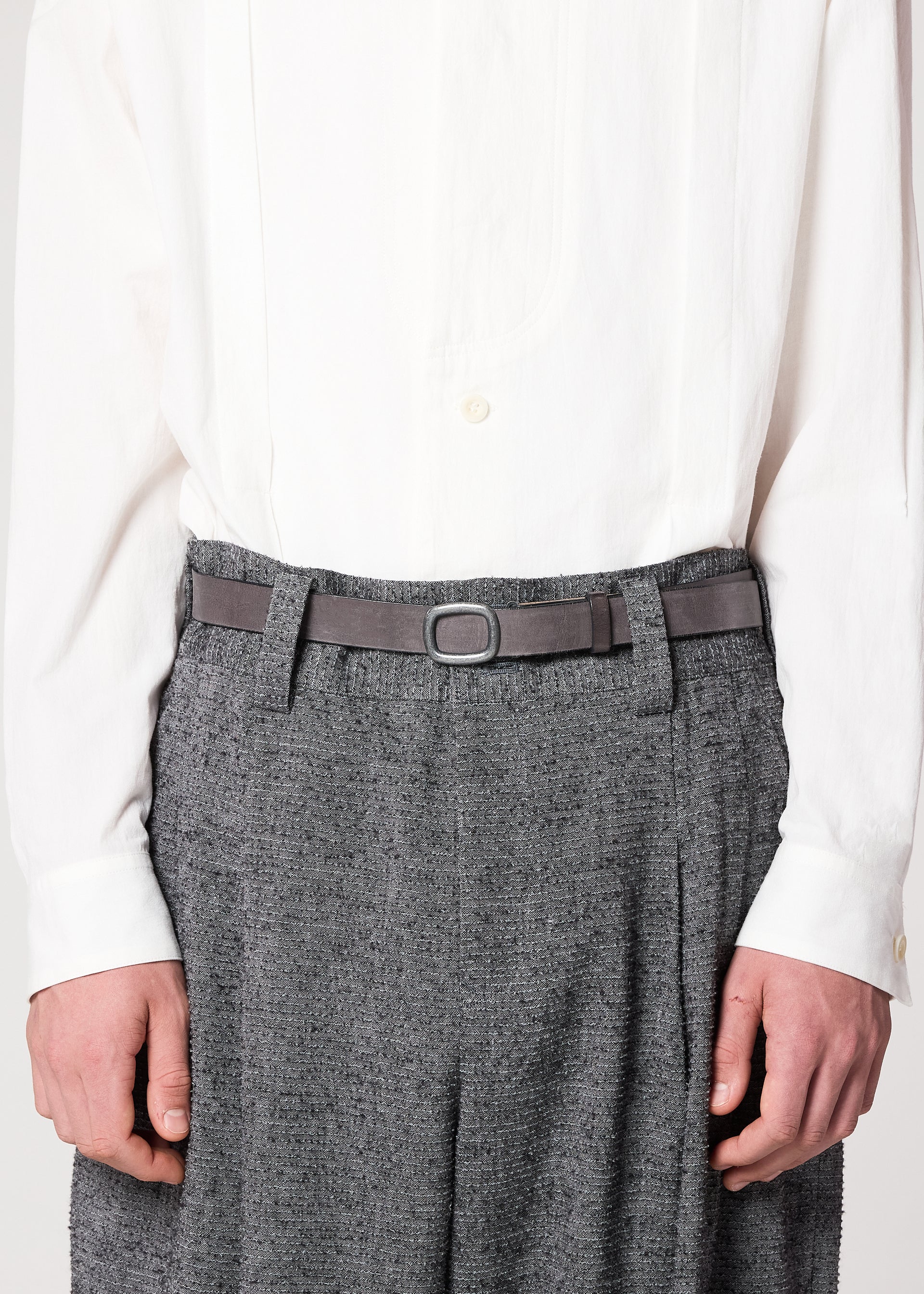 Zoomed view A person wearing IM MEN’s OVAL one-size-fits-most natural leather belt with an adjustable buckle, styled over gray textured trousers and a white button-up shirt. The photo is cropped at the shoulders and mid-thigh, hands at their sides.