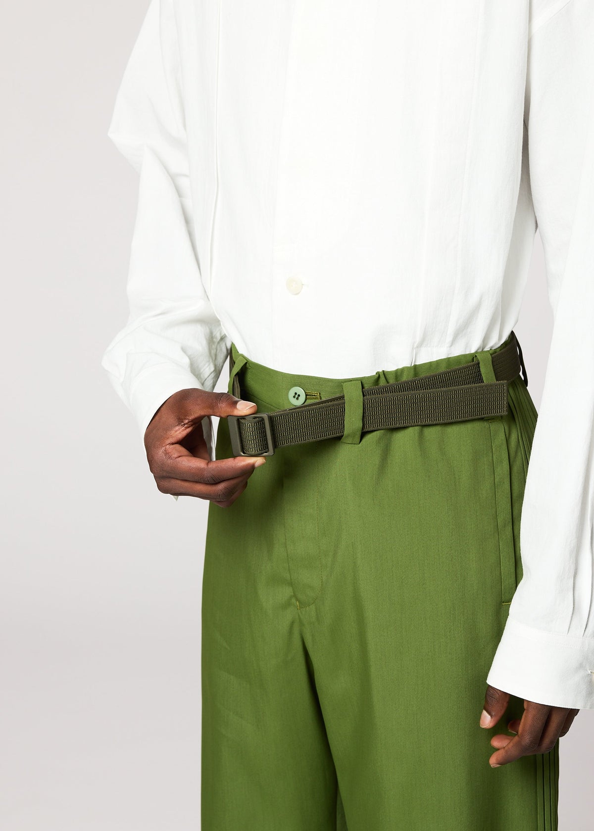 A person in a white button-up and high-waisted green pants fastens the olive-green FRICTION BELT by IM MEN. The image is cropped to display only their torso and hands against a plain background. view 6 of 6