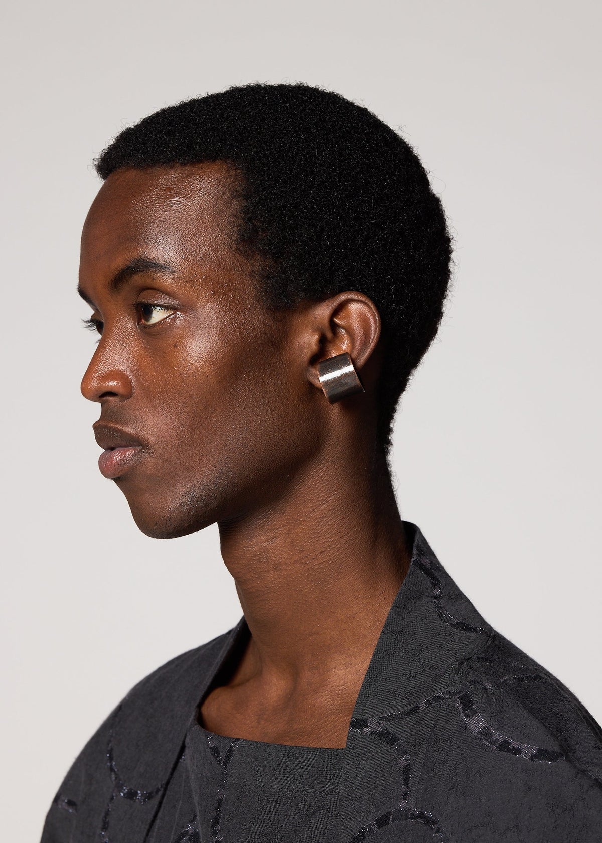 A person with short curly hair wears IM MEN's FASTEN RING, a large rectangular metallic ear cuff on their left ear, paired with a textured black top and set against a plain light background. view 3 of 4