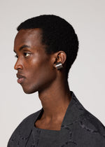 A person with short curly hair wears IM MEN's FASTEN RING, a large rectangular metallic ear cuff on their left ear, paired with a textured black top and set against a plain light background.