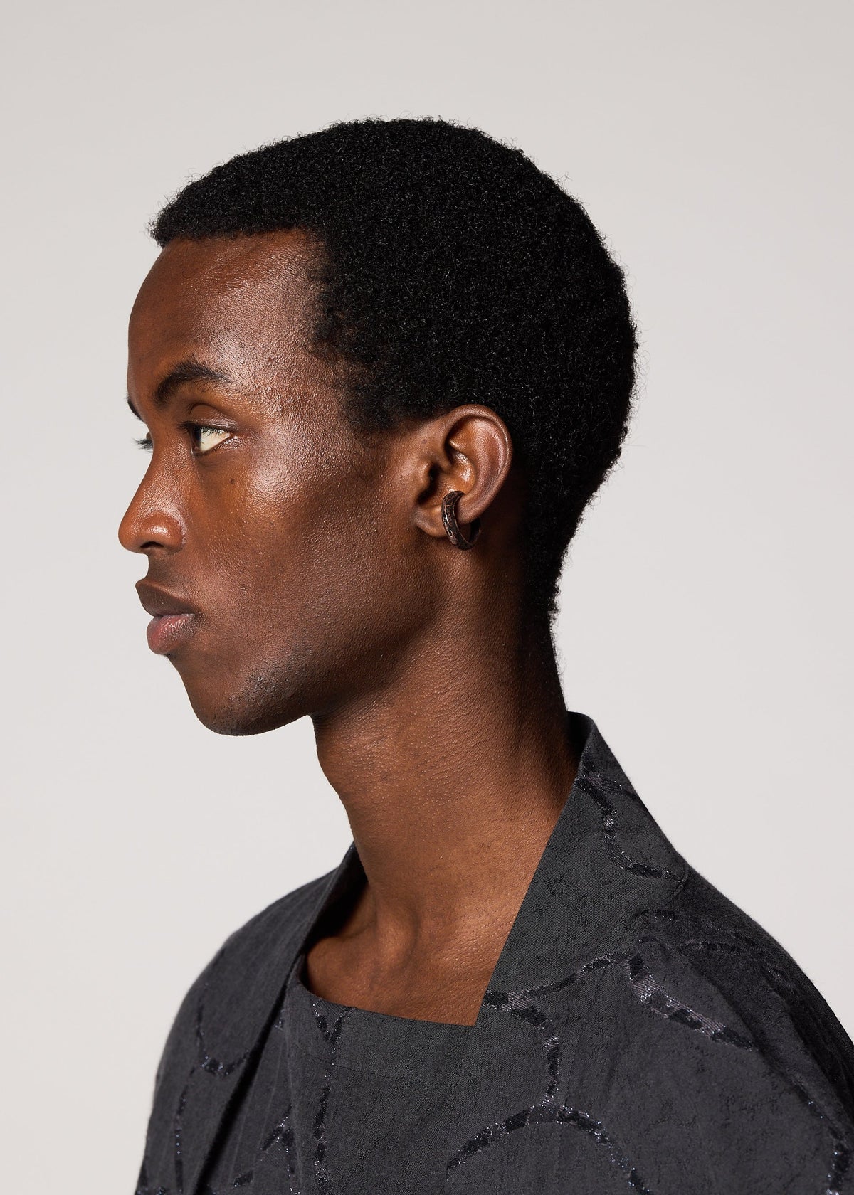 A young person with short, curly black hair is shown in profile facing left, wearing a dark patterned garment, a small hoop earring, and the elegant FASTEN RING by IM MEN. The background is plain and light-colored. view 3 of 4