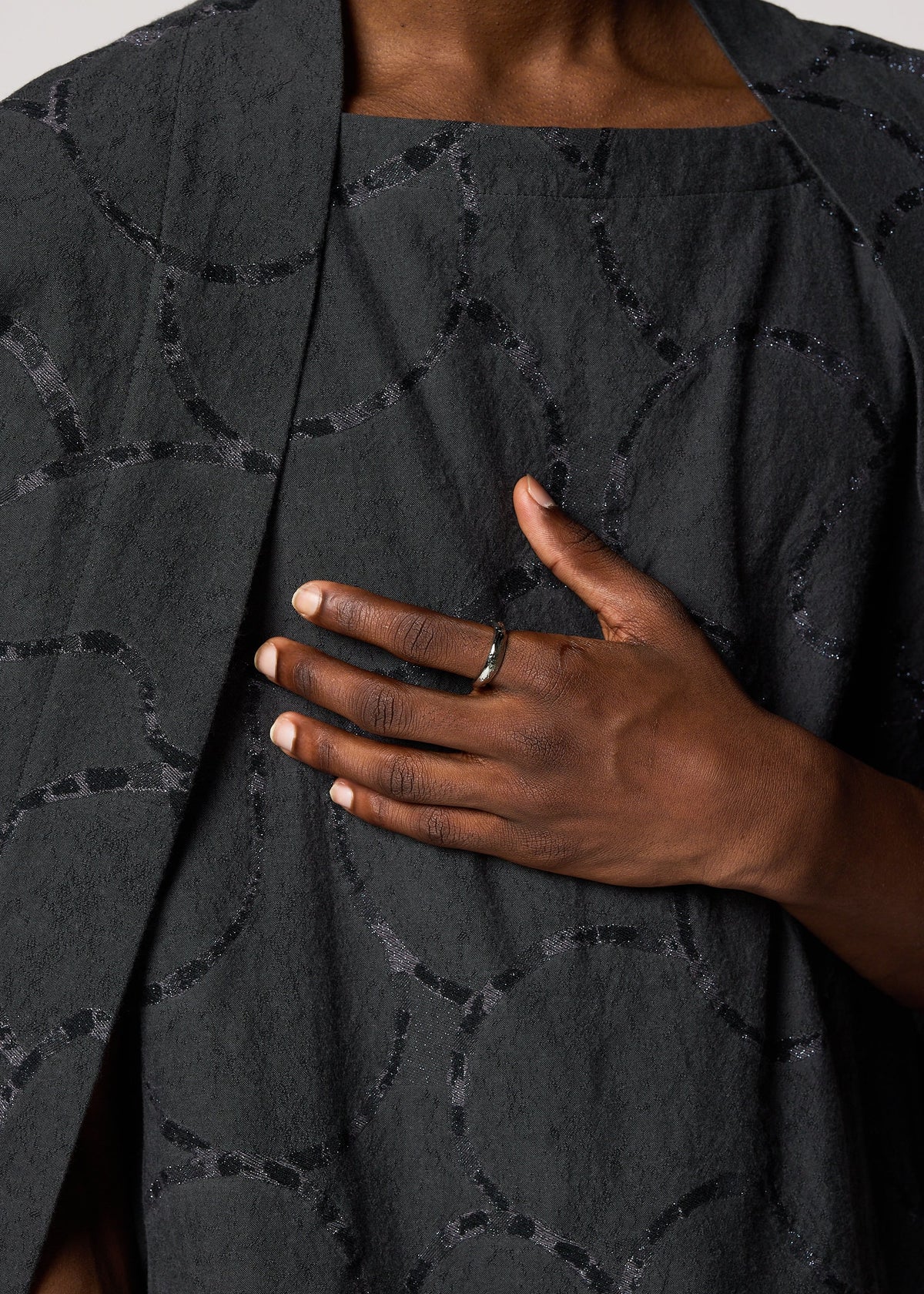A person with neatly manicured nails wears the IM MEN FASTEN RING, a simple ceramic accessory, on their finger while resting their hand on their chest over a textured dark gray outfit with a subtle circular pattern. view 4 of 4