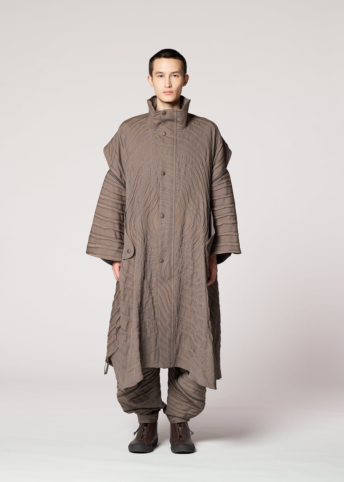 A person faces forward in IM MEN's ENGRAVE set: a loose taupe jacquard coat with wave patterns and wide sleeves, paired with matching quilted pants and dark shoes. The three-dimensional textile adds structure. The background is light gray. view 5 of 8