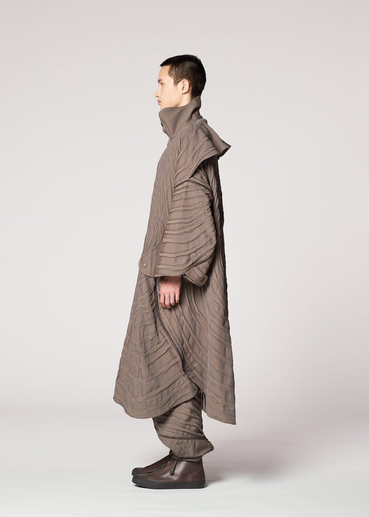 A person in profile models IM MEN’s ENGRAVE: a jacquard coat with a subtle wave pattern and matching pants. The oversized silhouette, high collar, and layered look stand out against a plain, light background. view 6 of 8