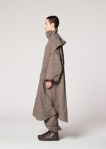 A person in profile models IM MEN’s ENGRAVE: a jacquard coat with a subtle wave pattern and matching pants. The oversized silhouette, high collar, and layered look stand out against a plain, light background.