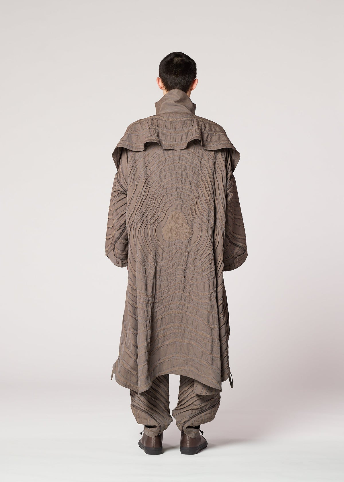A person faces away, wearing IM MEN's ENGRAVE: a long taupe jacquard coat with a high collar and wave patterns, paired with matching pants and shoes. The 3D textile stands out against the light background. view 7 of 8