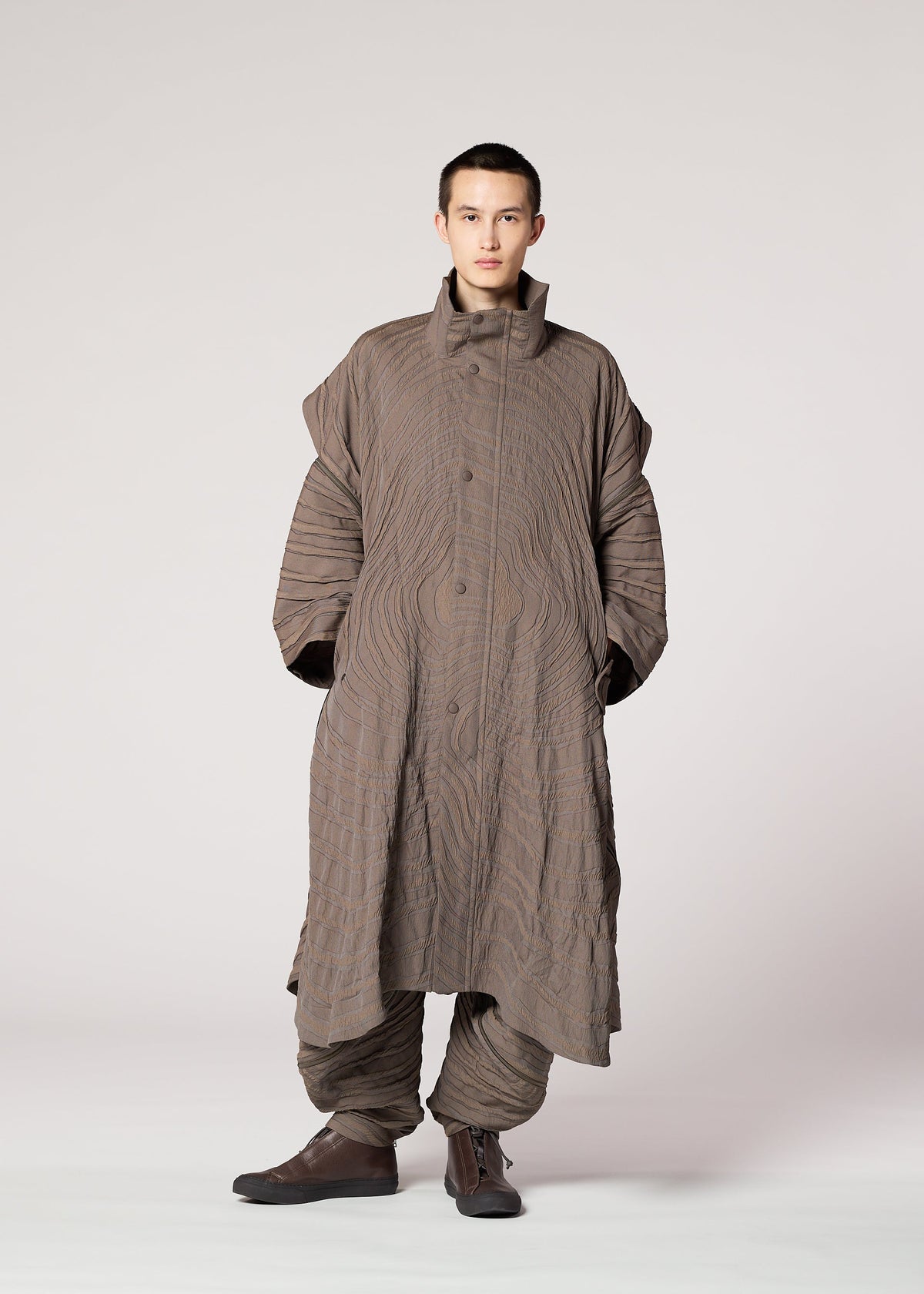A person stands facing forward in IM MEN's ENGRAVE: an oversized brown jacquard coat with a wavy 3D pattern, high collar, voluminous sleeves, and matching loose pants, set against a plain light gray background. view 8 of 8