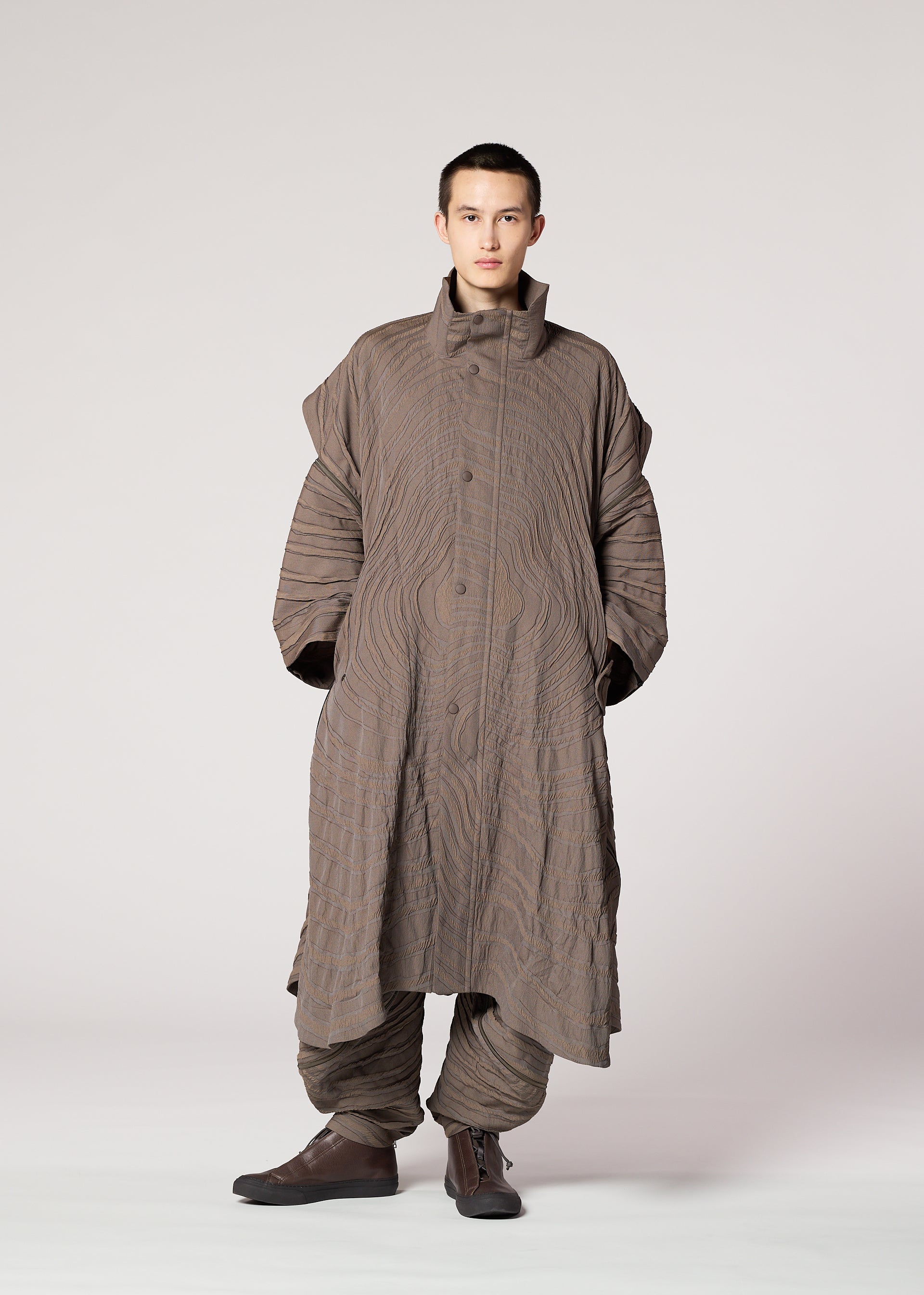 Zoomed view A person stands facing forward in IM MEN's ENGRAVE: an oversized brown jacquard coat with a wavy 3D pattern, high collar, voluminous sleeves, and matching loose pants, set against a plain light gray background.