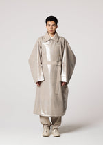 A person stands against a plain background, dressed in a Shoji Kamoda–inspired shiny metallic beige oversized trench coat with wide sleeves, beige pants, and completes the look with IM MEN GINTO FLAT light-colored shoes.