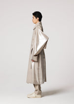 A person in profile wears the IM MEN GINTO FLAT: a Shoji Kamoda-inspired, long, shiny beige plant-based polyester coat with oversized sleeves, matching pants and shoes, all set against a plain light gray background.