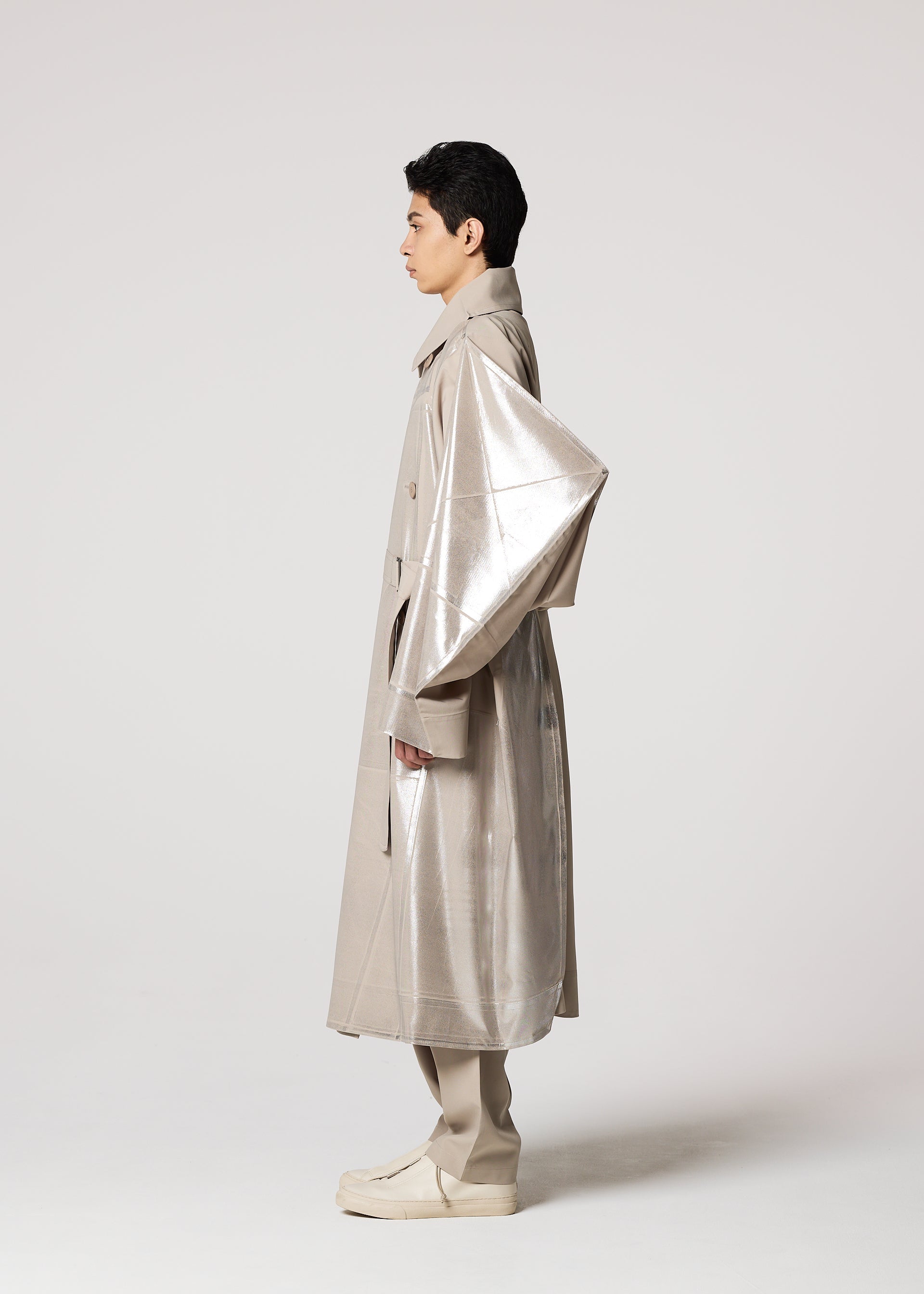 Zoomed view A person in profile wears the IM MEN GINTO FLAT: a Shoji Kamoda-inspired, long, shiny beige plant-based polyester coat with oversized sleeves, matching pants and shoes, all set against a plain light gray background.