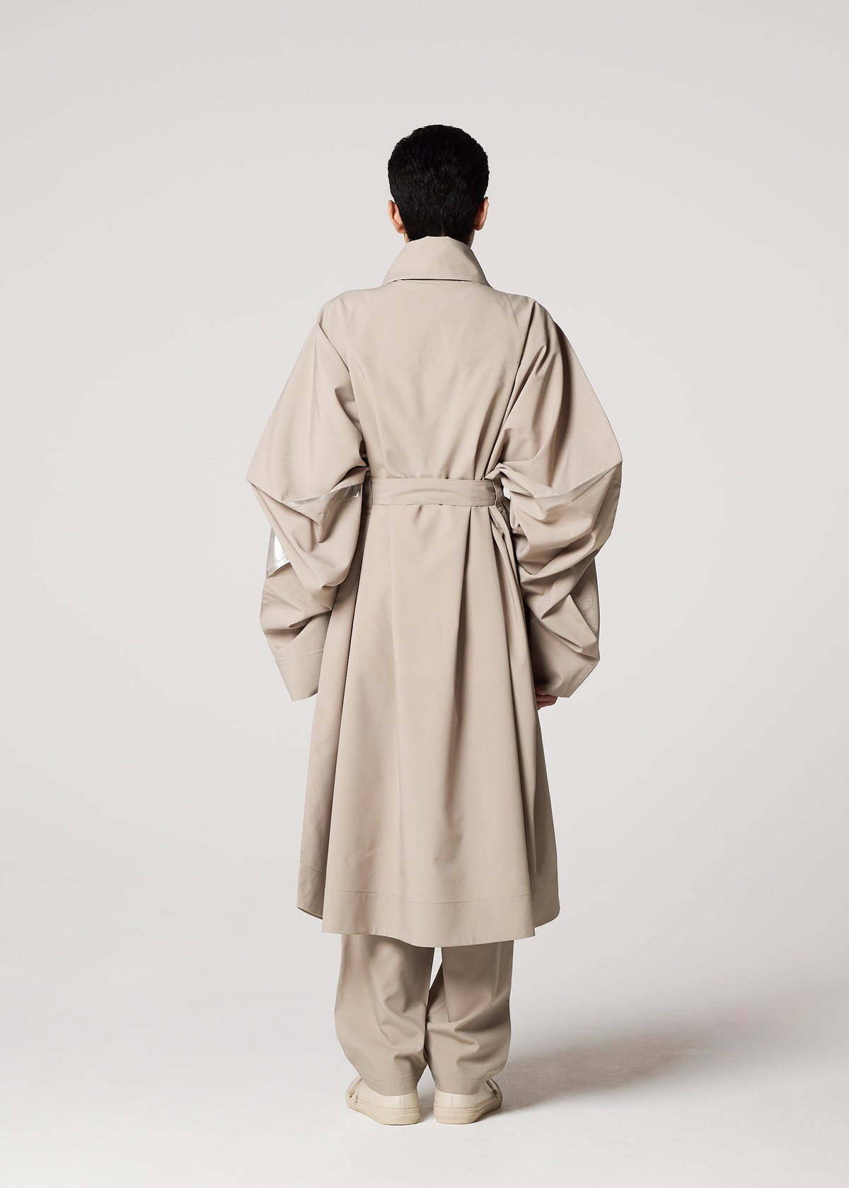 A person stands facing away in the GINTO FLAT by IM MEN—a Shoji Kamoda inspired beige trench coat with oversized sleeves and matching wide-leg pants—set against a plain, light-colored background. view 7 of 9