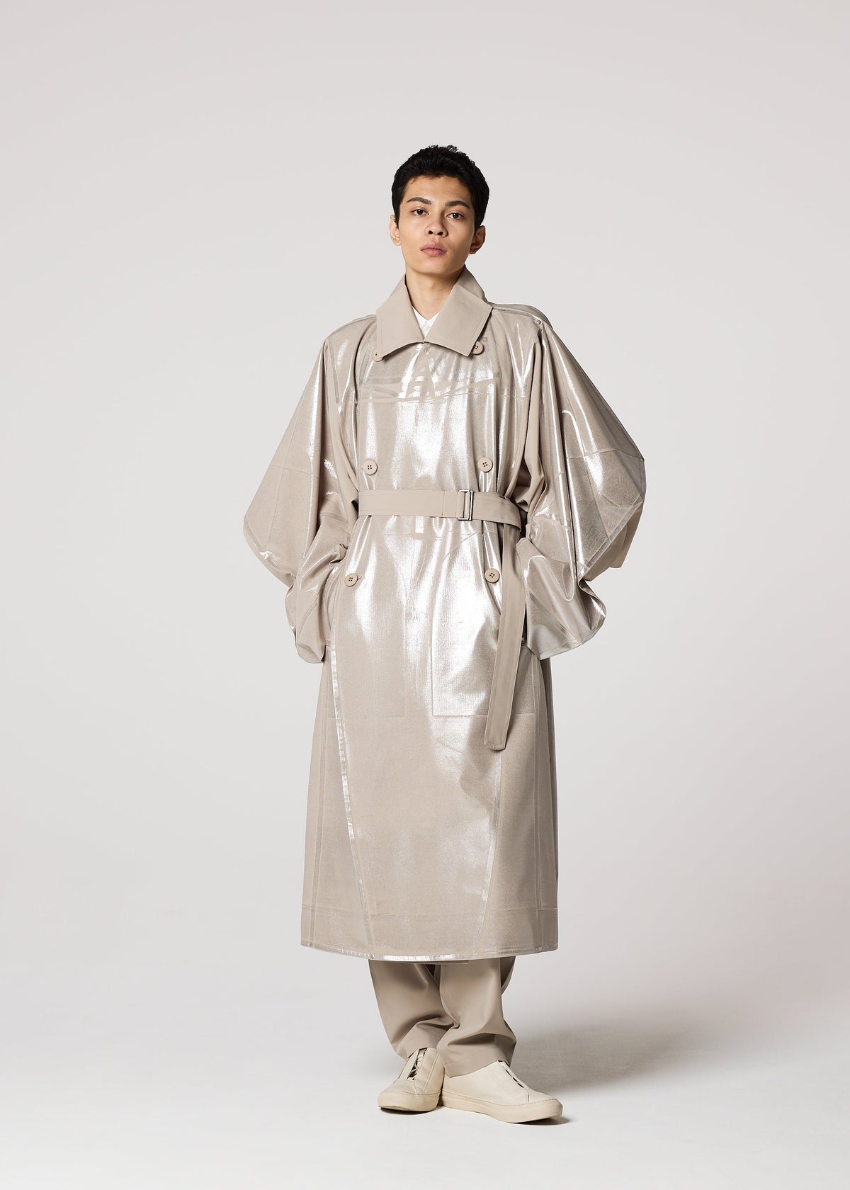 A person stands facing forward in a shiny, Shoji Kamoda-inspired light beige trench coat with wide sleeves, beige pants, and IM MEN GINTO FLAT cream-colored shoes against a plain light background. view 8 of 9