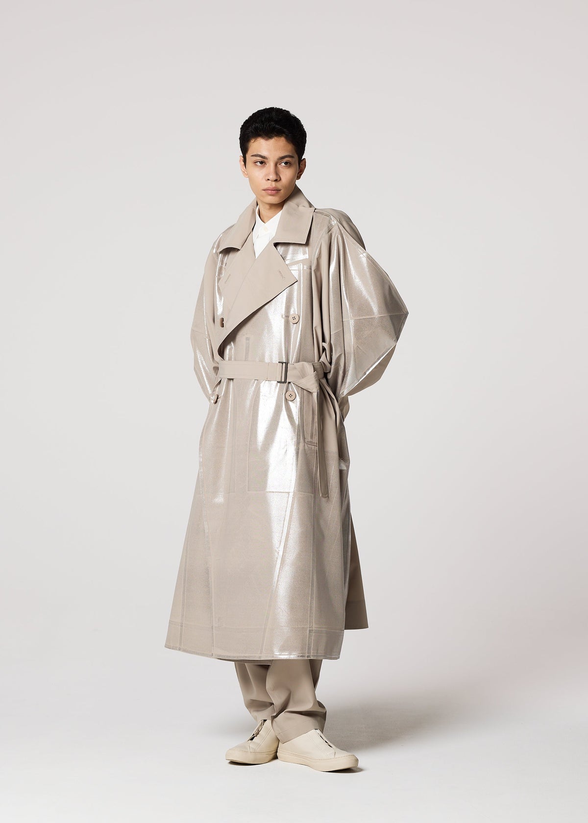 Against a plain light background, a person wears an oversized, shiny plant-based polyester GINTO FLAT coat with wide lapels and matching pants by IM MEN. Light-colored shoes complete the look as they stand hands behind their back, gazing to the side. view 9 of 9