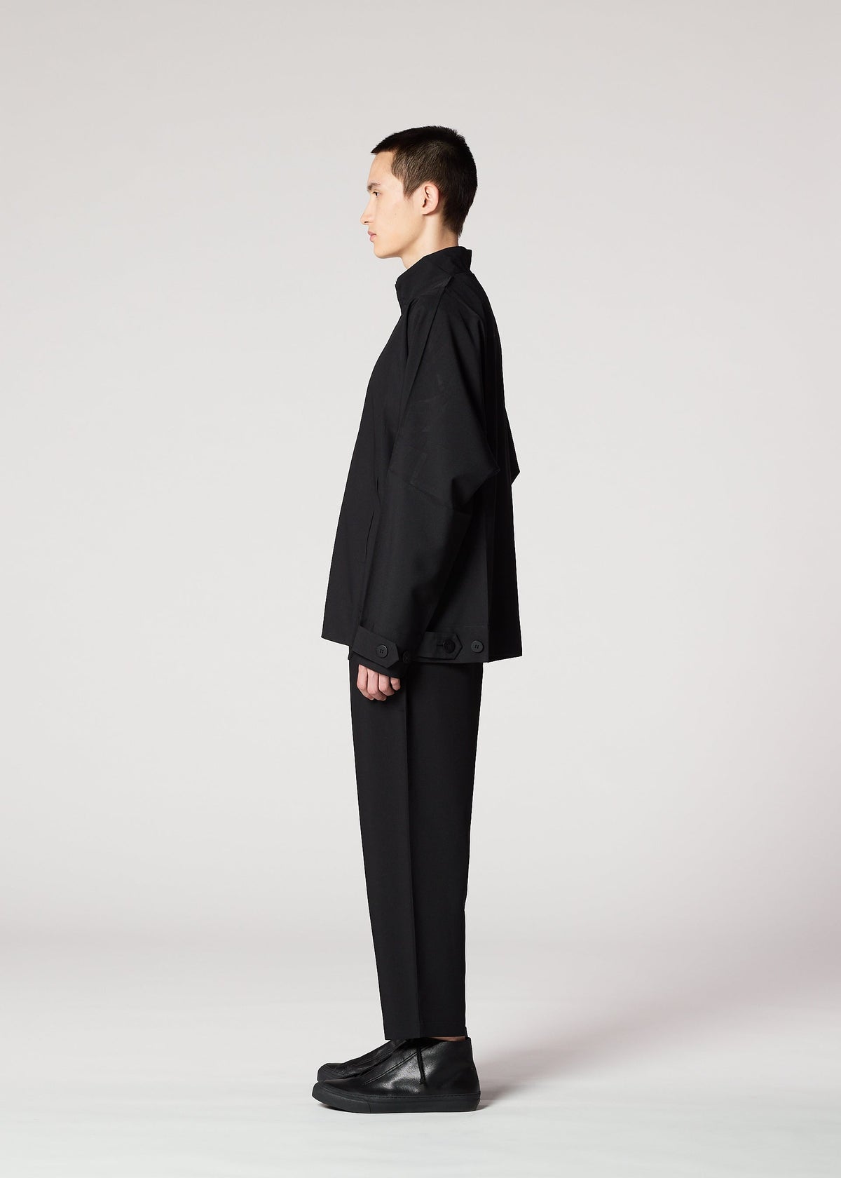 A person with short dark hair stands in profile against a plain light background, wearing IM MEN’s FLAT BLACK loose-fitting, wrinkle-resistant jacket, paired with black pants and black shoes. view 11 of 14