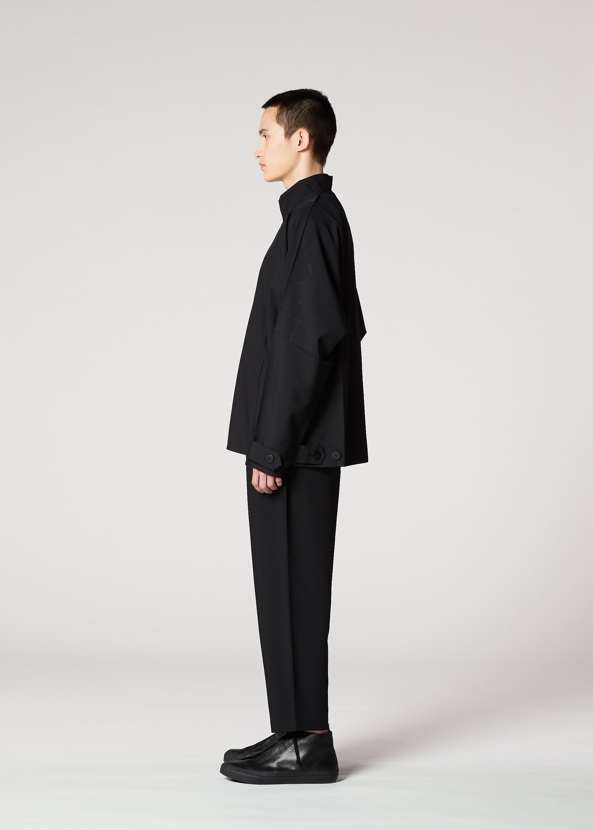 Zoomed view A person with short dark hair stands in profile against a plain light background, wearing IM MEN’s FLAT BLACK loose-fitting, wrinkle-resistant jacket, paired with black pants and black shoes.
