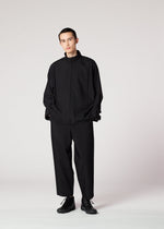 A person stands against a plain light background, hands in pockets and a neutral expression, wearing the IM MEN FLAT BLACK loose-fitting, wrinkle-resistant jacket with wide black trousers and black shoes.