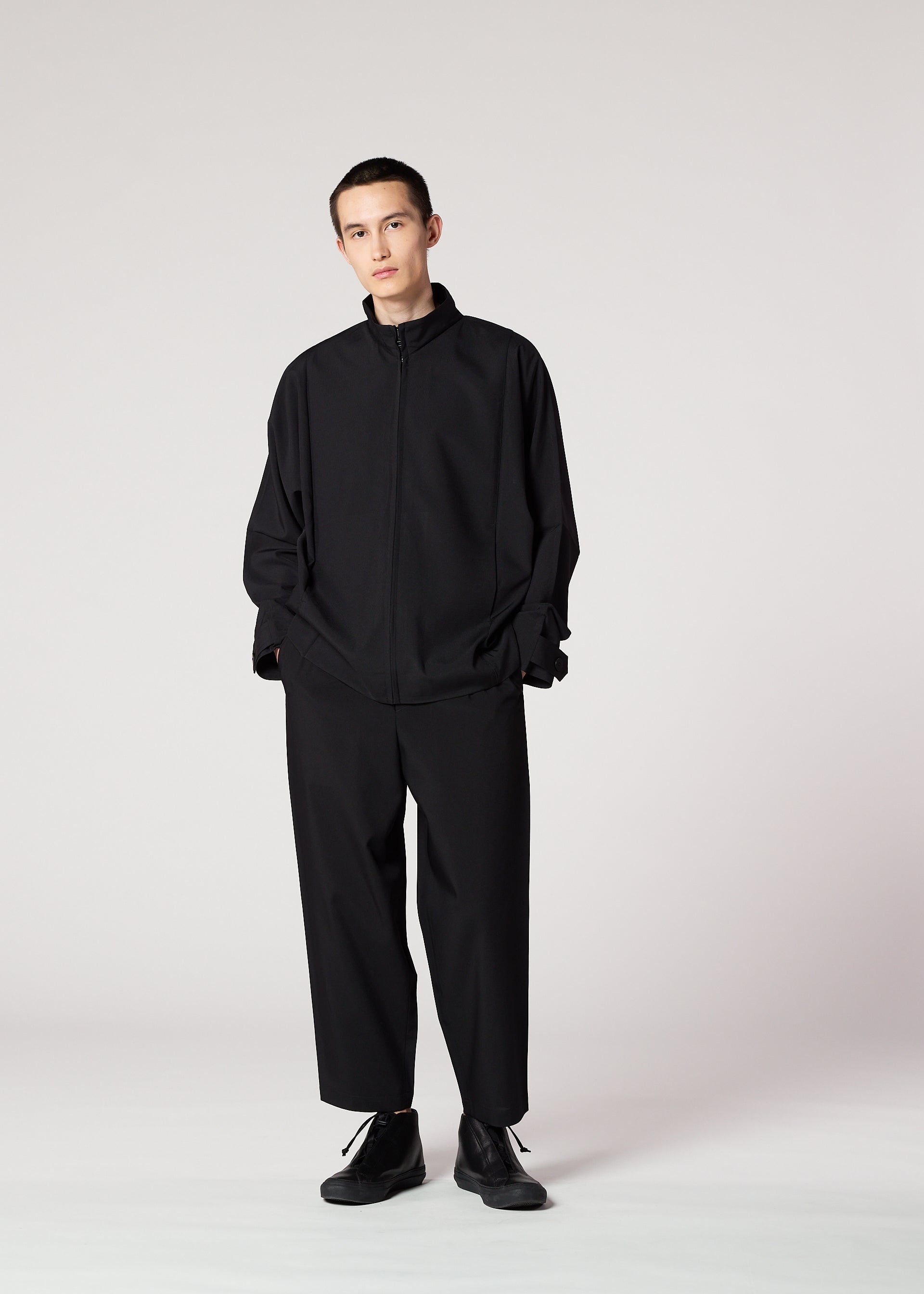 Zoomed view A person stands against a plain light background, hands in pockets and a neutral expression, wearing the IM MEN FLAT BLACK loose-fitting, wrinkle-resistant jacket with wide black trousers and black shoes.