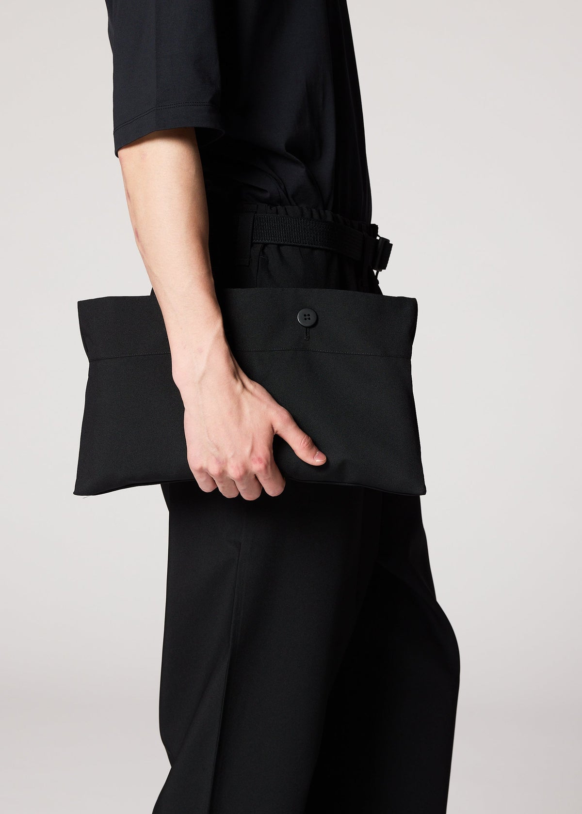 A person dressed in black holds the IM MEN FLAT BLACK rectangular clutch with a button closure at their side, ideal for carrying essentials like a compact portable jacket, against a plain light background. view 14 of 14