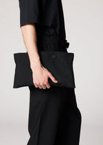 A person dressed in black holds the IM MEN FLAT BLACK rectangular clutch with a button closure at their side, ideal for carrying essentials like a compact portable jacket, against a plain light background.