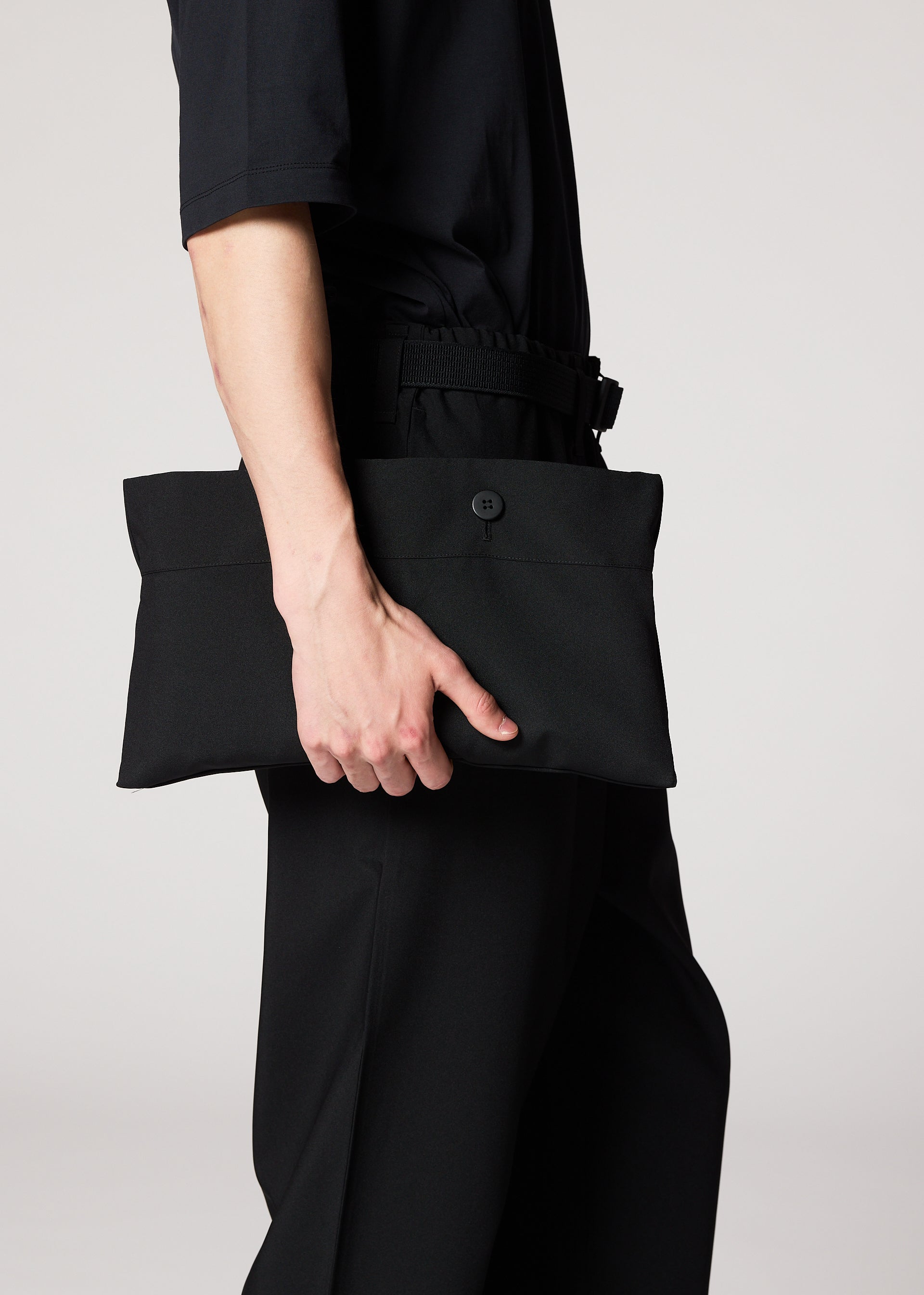 Zoomed view A person dressed in black holds the IM MEN FLAT BLACK rectangular clutch with a button closure at their side, ideal for carrying essentials like a compact portable jacket, against a plain light background.