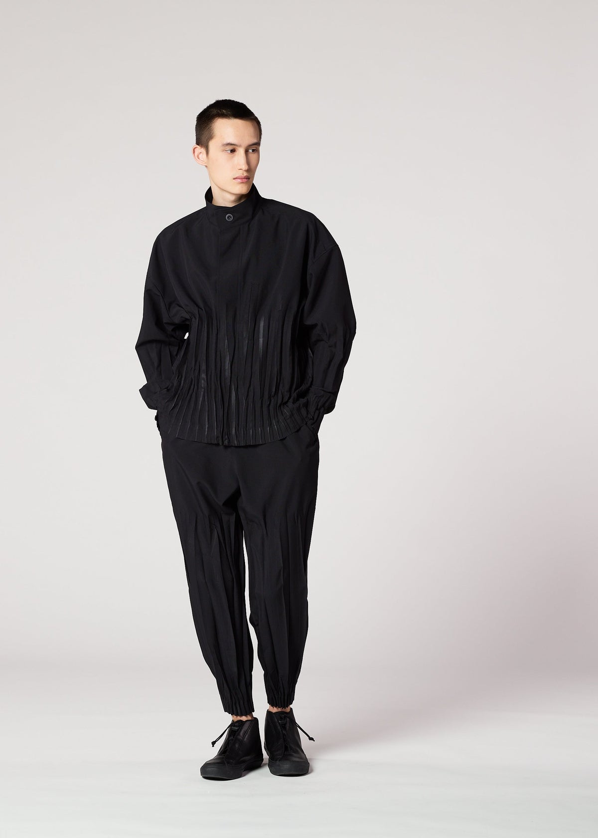 A person stands against a plain background, wearing the IM MEN WAFT BLACK set: a loose-fitting black jacket with pleated details and matching wrinkle-resistant pants with gathered ankles, paired with black shoes. view 14 of 15