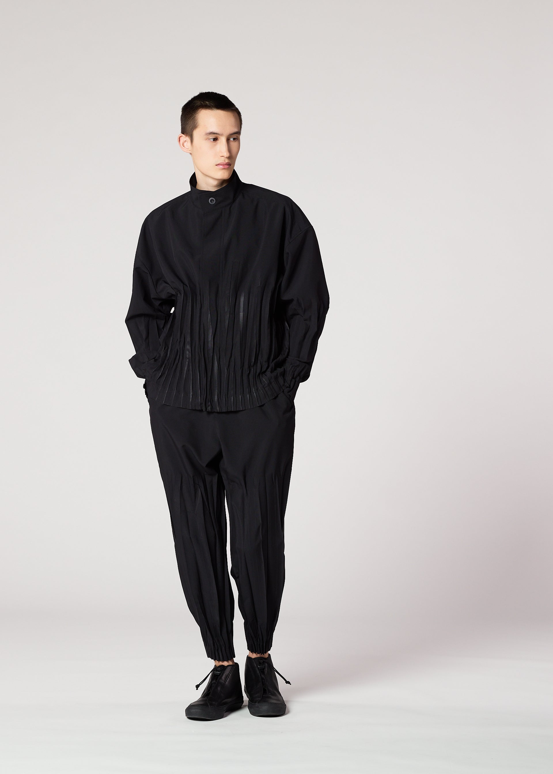 Zoomed view A person stands against a plain background, wearing the IM MEN WAFT BLACK set: a loose-fitting black jacket with pleated details and matching wrinkle-resistant pants with gathered ankles, paired with black shoes.