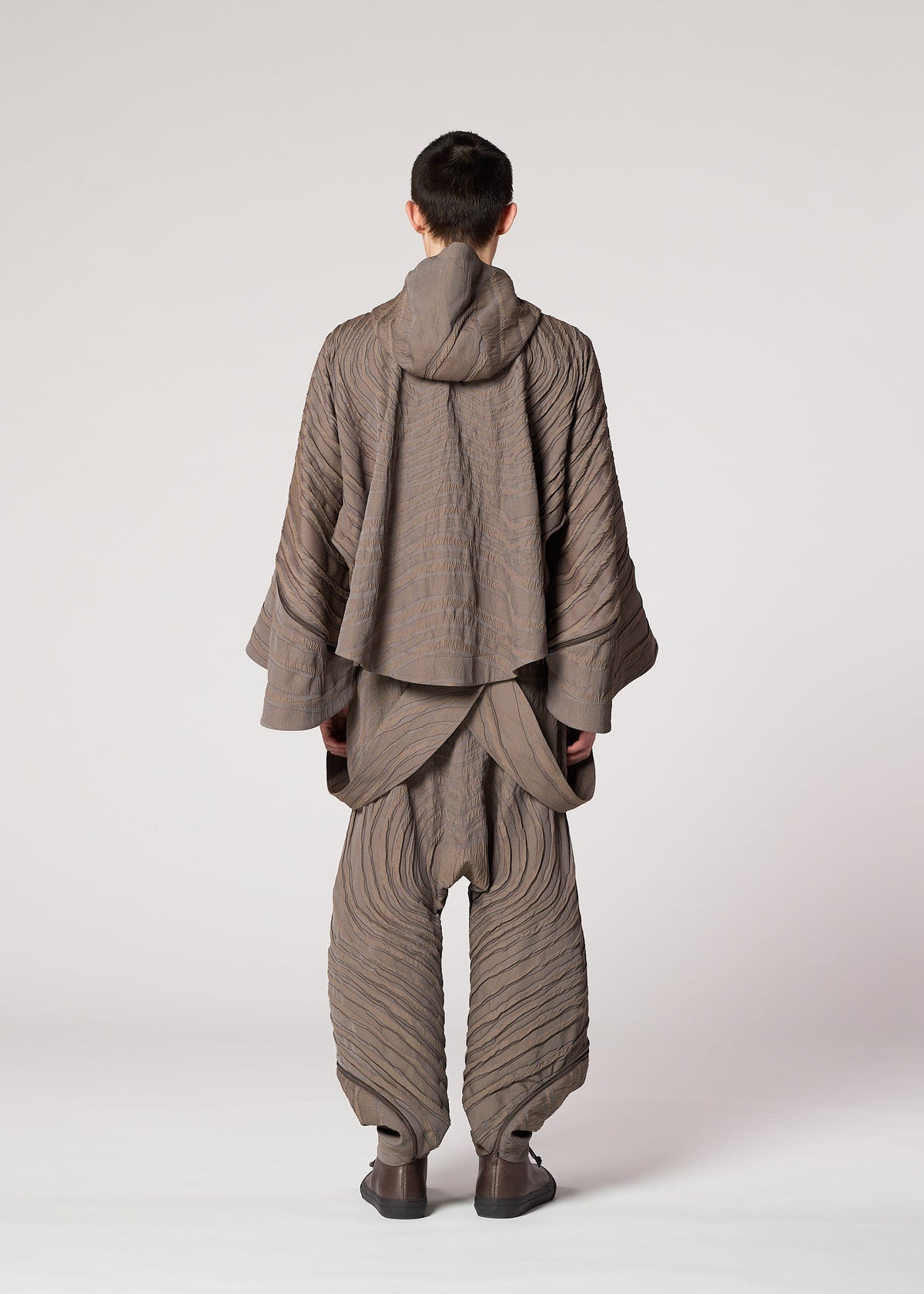 A person faces away, wearing IM MEN's ENGRAVE: a Shoji Kamoda-inspired taupe outfit with wide pants and a hooded, layered top. The textured design evokes three-dimensional wave patterns against a light background. view 7 of 9